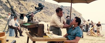 Movie still from “Babylon” (2022), directed by Damien Chazelle – An old man getting his face shaved by an older man on the beach; Medium shot, Over the shoulder angle