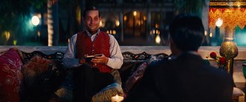 Movie still from “Babylon” (2022), directed by Damien Chazelle – A man sitting on top of a couch while holding a cell phone; Medium shot, Over the shoulder angle