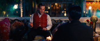 Movie still from “Babylon” (2022), directed by Damien Chazelle – A man sitting on top of a couch next to a candle; Medium shot, Over the shoulder angle