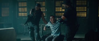 Movie still from “The Expendables 2” (2012), directed by Simon West – A group of men in a room with a man sitting on the ground; Medium shot, Over the shoulder angle