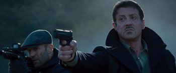 Movie still from “The Expendables 2” (2012), directed by Simon West – Two men in black jackets holding a black gun; Close Up shot, Low angle