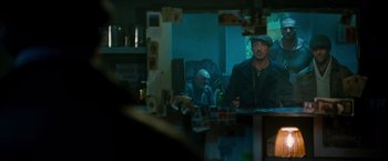 Movie still from “The Expendables 2” (2012), directed by Simon West – Two men sitting at a table with a bottle of beer; Medium shot, Over the shoulder angle
