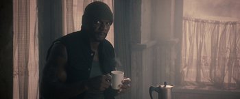 Movie still from “The Expendables 2” (2012), directed by Simon West – A man holding a cup of coffee while sitting in a room; Medium shot, Over the shoulder angle
