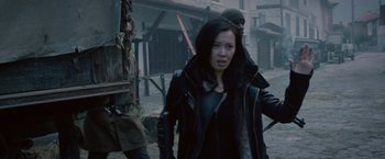 Movie still from “The Expendables 2” (2012), directed by Simon West – A woman wearing a black jacket walking down a street; Medium shot, Over the shoulder angle