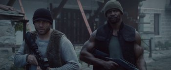 Movie still from “The Expendables 2” (2012), directed by Simon West – A man holding a gun next to another man; Medium shot, Over the shoulder angle