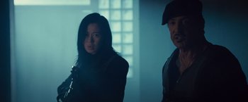 Movie still from “The Expendables 2” (2012), directed by Simon West – A woman standing next to a man in a dark room; Medium shot, Low angle