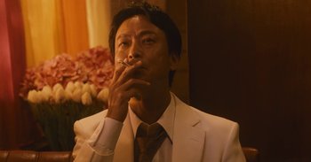 Movie still from “The Forest of Love” (2019), directed by Sion Sono – A man in a suit smoking a cigarette in front of flowers; Close Up shot, Low angle