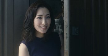 Movie still from “The Forest of Love” (2019), directed by Sion Sono – A woman standing in front of a door smiling for the camera; Close Up shot, Over the shoulder angle