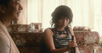 Movie still from “The Forest of Love” (2019), directed by Sion Sono – A woman holding a bottle of beer while sitting on a couch; Medium shot, High angle