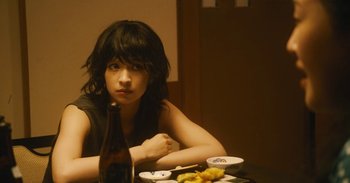 Movie still from “The Forest of Love” (2019), directed by Sion Sono – A woman sitting at a table with a bottle of wine; Close Up shot, Over the shoulder angle