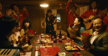 Movie still from “The Forest of Love” (2019), directed by Sion Sono – A group of people sitting around a table; Wide shot, High angle