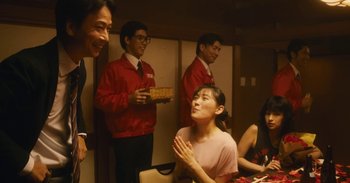 Movie still from “The Forest of Love” (2019), directed by Sion Sono – A group of people standing around a table; Medium shot, Over the shoulder angle