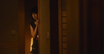 Movie still from “The Forest of Love” (2019), directed by Sion Sono – A woman standing in front of a door in a room; Medium shot, Over the shoulder angle