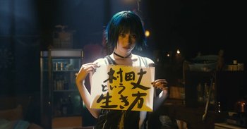 Movie still from “The Forest of Love” (2019), directed by Sion Sono – A woman holding up a sign in a dark room; Medium shot, Low angle