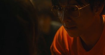 Movie still from “The Forest of Love” (2019), directed by Sion Sono – A person wearing glasses sitting down in a room; Close Up shot, Low angle