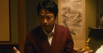 Movie still from “The Forest of Love” (2019), directed by Sion Sono – A man in a red suit is standing in front of a painting; Close Up shot, Low angle
