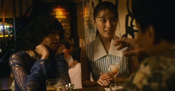 Movie still from “The Forest of Love” (2019), directed by Sion Sono – A woman sitting at a table with a glass of wine; Medium shot, Over the shoulder angle