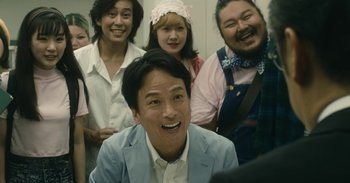 Movie still from “The Forest of Love” (2019), directed by Sion Sono – A group of people standing around each other smiling for the camera; Medium shot, Over the shoulder angle