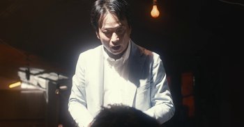 Movie still from “The Forest of Love” (2019), directed by Sion Sono – A man in a white suit is holding something in his hands; Close Up shot, Low angle