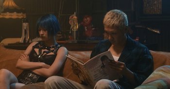 Movie still from “The Forest of Love” (2019), directed by Sion Sono – A man and a woman sitting on a couch looking at a magazine; Medium shot, Low angle