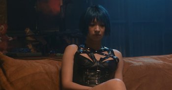 Movie still from “The Forest of Love” (2019), directed by Sion Sono – A woman sitting on top of a brown couch wearing a black corset; Medium shot, Over the shoulder angle