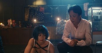 Movie still from “The Forest of Love” (2019), directed by Sion Sono – A woman sitting on the ground next to a man; Medium shot, Over the shoulder angle