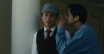 Movie still from “The Forest of Love” (2019), directed by Sion Sono – A man whispering into a man's ear while standing next to another man; Close Up shot, Over the shoulder angle