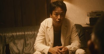 Movie still from “The Forest of Love” (2019), directed by Sion Sono – A man sitting on top of a couch in a room; Medium shot, Low angle