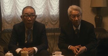 Movie still from “The Forest of Love” (2019), directed by Sion Sono – Two men in business suits sitting at a table; Medium shot, Low angle