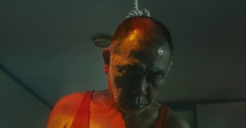 Movie still from “The Forest of Love” (2019), directed by Sion Sono – A man in a red tank top with a rope hanging from his head; Close Up shot, Low angle