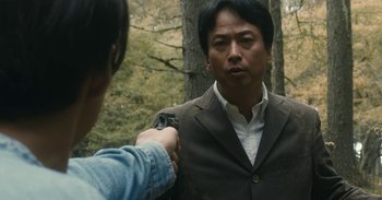 Movie still from “The Forest of Love” (2019), directed by Sion Sono – A man holding a gun pointed at another man in a forest; Medium shot, Over the shoulder angle