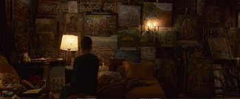 Movie still from “The Glass Castle” (2017), directed by Destin Daniel Cretton – A person sitting on a bed in a room filled with paintings; Wide shot, Overhead angle