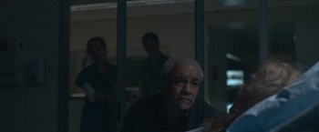 Movie still from “The Good Nurse” (2022), directed by Tobias Lindholm – An older man sitting in front of a glass wall; Medium shot, Over the shoulder angle
