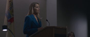 Movie still from “The Good Nurse” (2022), directed by Tobias Lindholm – A woman in a blue suit standing at a podium with a microphone; Medium shot, Over the shoulder angle