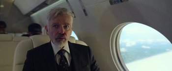 Movie still from “The Gray Man” (2022), directed by Joe Russo – An older man wearing a suit and tie on an airplane; Close Up shot, Over the shoulder angle
