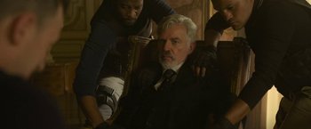 Movie still from “The Gray Man” (2022), directed by Joe Russo – A man sitting in a chair with another man standing behind him; Medium shot, Over the shoulder angle