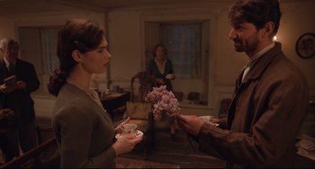 Movie still from “The Guernsey Literary and Potato Peel Pie Society” (2018), directed by Mike Newell – A man and a woman holding flowers in a living room; Medium shot, Over the shoulder angle