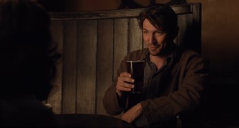 Movie still from “The Guernsey Literary and Potato Peel Pie Society” (2018), directed by Mike Newell – A man sitting at a table holding a glass of beer; Medium shot, Over the shoulder angle