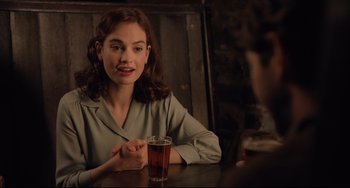 Movie still from “The Guernsey Literary and Potato Peel Pie Society” (2018), directed by Mike Newell – A woman sitting at a table with a glass of beer; Medium shot, Over the shoulder angle