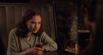 Movie still from “The Guernsey Literary and Potato Peel Pie Society” (2018), directed by Mike Newell – A woman sitting at a table holding a glass of beer; Medium shot, Over the shoulder angle
