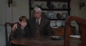 Movie still from “The Guernsey Literary and Potato Peel Pie Society” (2018), directed by Mike Newell – An older man sitting next to a young boy in front of plates; Medium shot, Over the shoulder angle