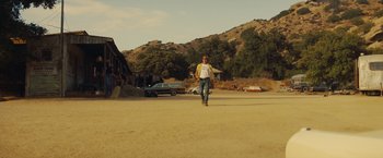 Movie still from “Once Upon a Time in Hollywood” (2019), directed by Quentin Tarantino – A man walking across a dirt road holding a skateboard; Wide shot, Low angle