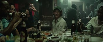 Movie still from “The Gunman” (2015), directed by Pierre Morel – A man sitting at a table with a bottle of beer; Medium shot, Over the shoulder angle