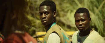 Movie still from “The Gunman” (2015), directed by Pierre Morel – A young man in a tan vest is standing in front of a tree; Close Up shot, Over the shoulder angle