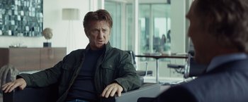 Movie still from “The Gunman” (2015), directed by Pierre Morel – A man sitting on a chair in a room; Medium shot, Over the shoulder angle