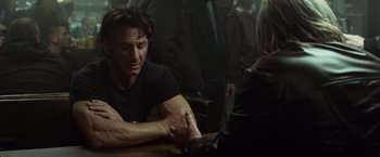 Movie still from “The Gunman” (2015), directed by Pierre Morel – A man sitting at a wooden table with a woman; Medium shot, Over the shoulder angle