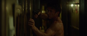 Movie still from “The Gunman” (2015), directed by Pierre Morel – A shirtless man holding a gun in a room; Medium shot, Low angle