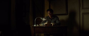 Movie still from “The Gunman” (2015), directed by Pierre Morel – A man sitting at a desk with a lamp on; Medium shot, Low angle