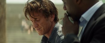 Movie still from “The Gunman” (2015), directed by Pierre Morel – A person sitting down with a cigarette in his mouth; Close Up shot, Over the shoulder angle
