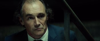 Movie still from “The Gunman” (2015), directed by Pierre Morel – An older man in a suit and tie looking at the camera; Close Up shot, Low angle